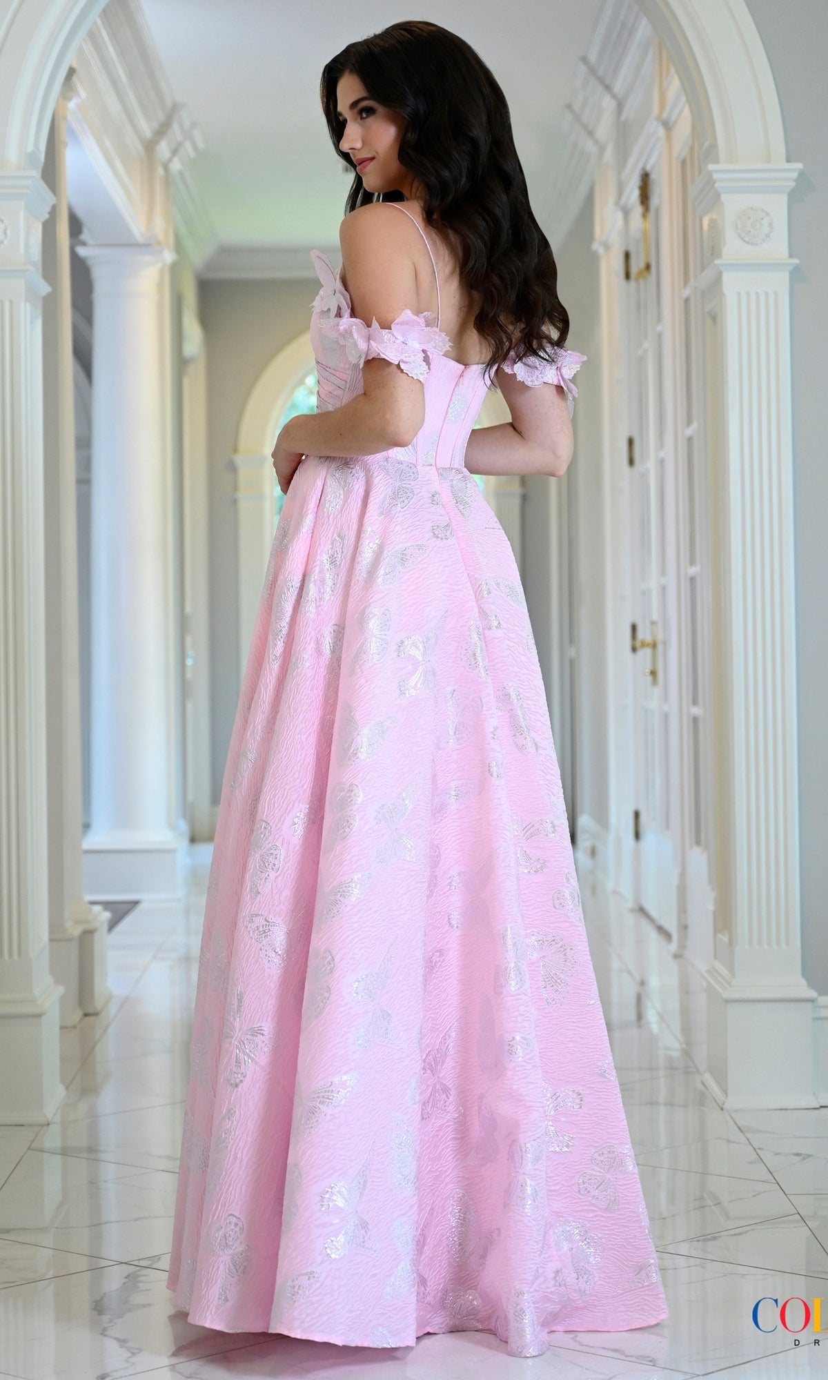A woman wears a floor-length pink jacquard Long Prom Dress: Colors Dress 4010 with off-the-shoulder sleeves and a silver floral pattern.